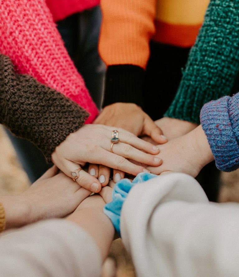 Hände von mehreren Personen, die sich in der Mitte übereinander legen, in bunten Pullovern.