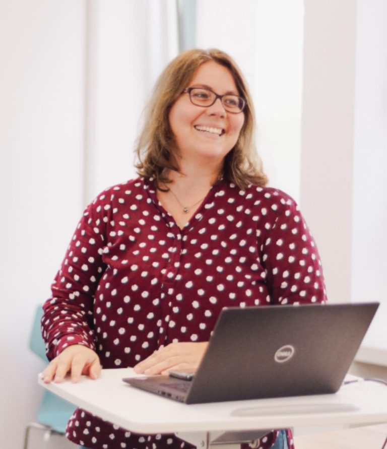 Lächelnde Frau mit Brille, steht an einem Tisch mit einem Laptop, trägt ein gepunktetes Hemd.