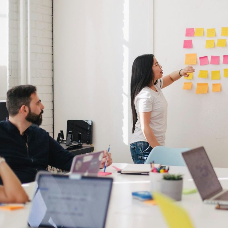 Eine Person erklärt ein Konzept an einer Wand mit Haftnotizen, andere hören aufmerksam zu.