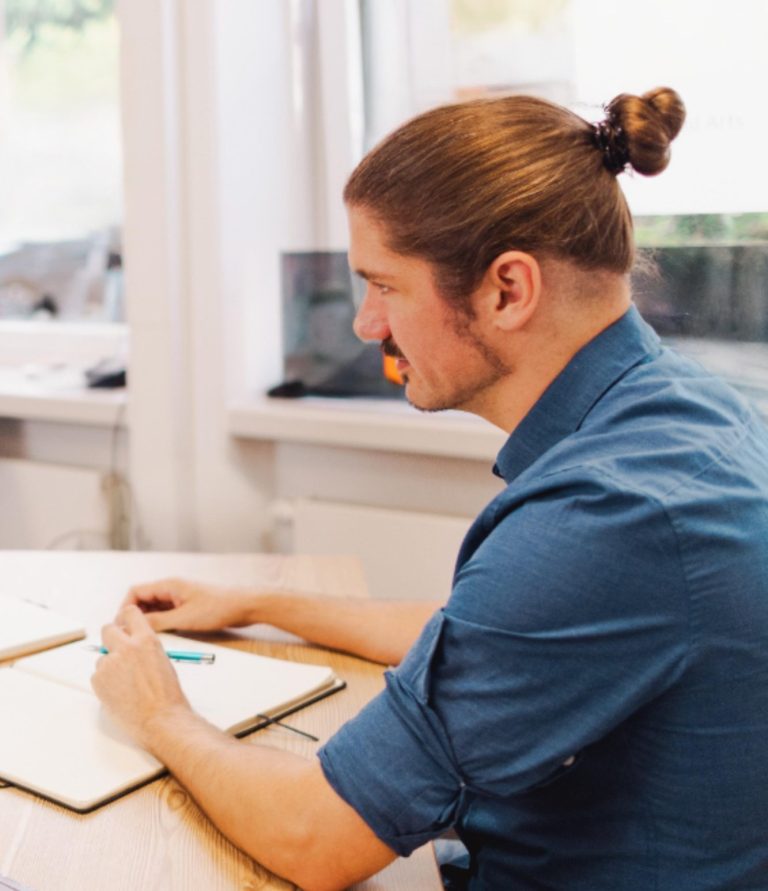 Mann mit Zopf, der an einem Tisch sitzt und in ein Notizbuch schreibt.