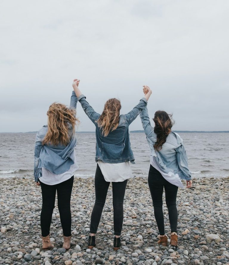 Drei Frauen stehen am Strand, heben die Arme in die Luft und schauen aufs Wasser.